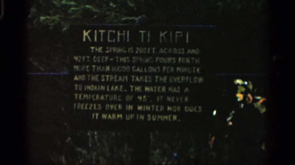 1960 Michigan 8mm frame showing a dark Kitchi Ti Kipi informational plaque with pale lettering and greenery behind it.