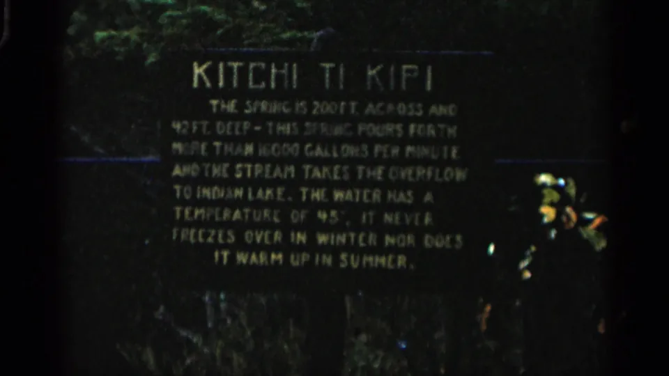 1960s 8mm home-movie frame of the Kitchi Ti Kipi spring plaque, softly exposed with visible grain and outdoor greenery.