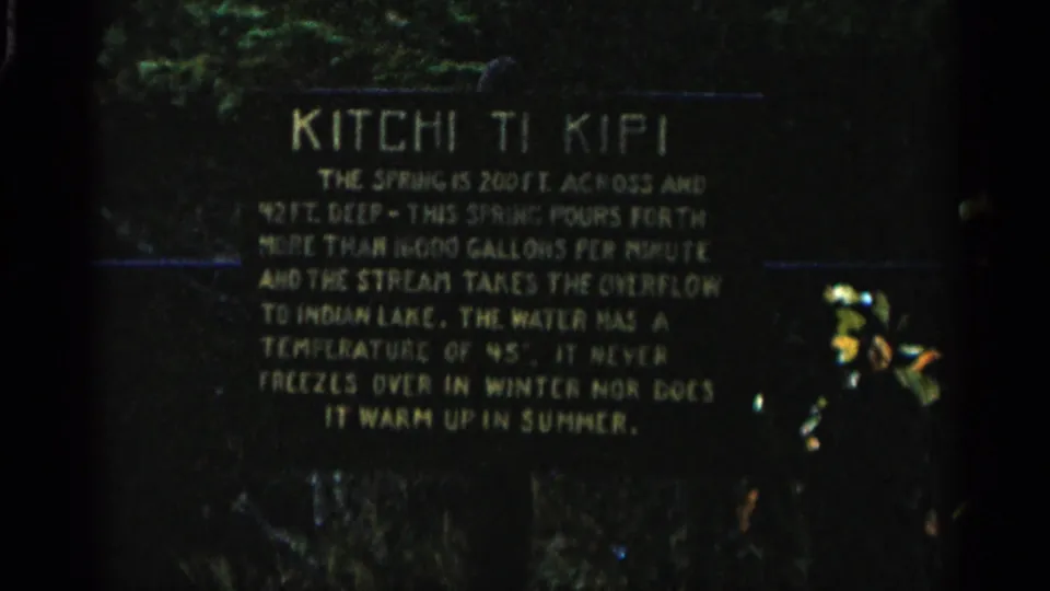 Michigan 8mm footage frame showing the Kitchi Ti Kipi informational sign, with pale text, dark board, and vegetation in the background.