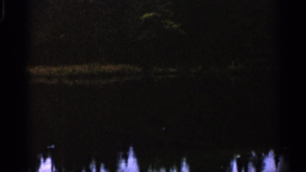 1960 Michigan 8mm frame showing dim water, soft shoreline reflections, and dark tree-like forms in low light.