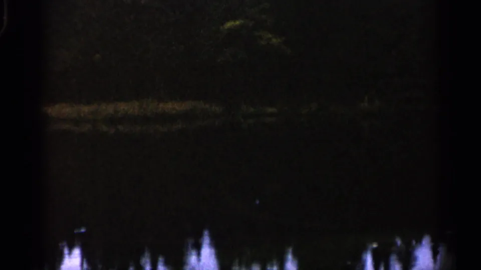 1960 Michigan 8mm frame showing dim water, soft shoreline reflections, and dark tree-like forms in low light.