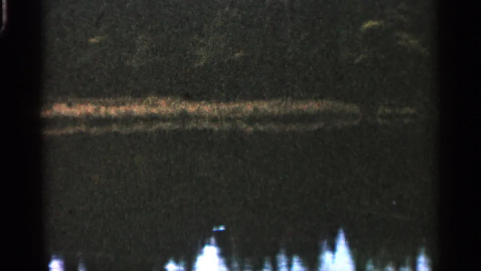 1960 Michigan 8mm frame showing dark water, a broad reflected band, and clustered pale shapes near the lower frame.