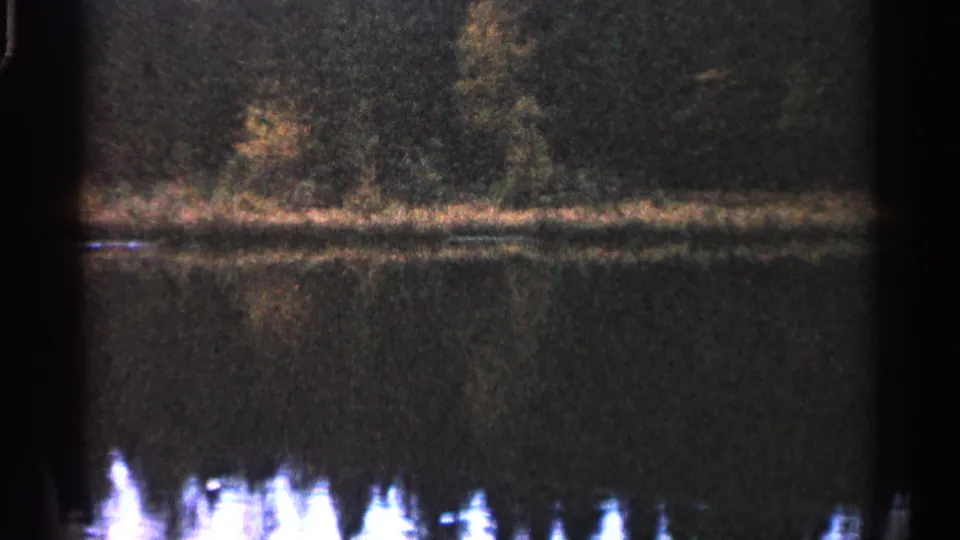 Grainy 8mm view of a Michigan lake, with trees along the bank and bright reflected highlights on the water.