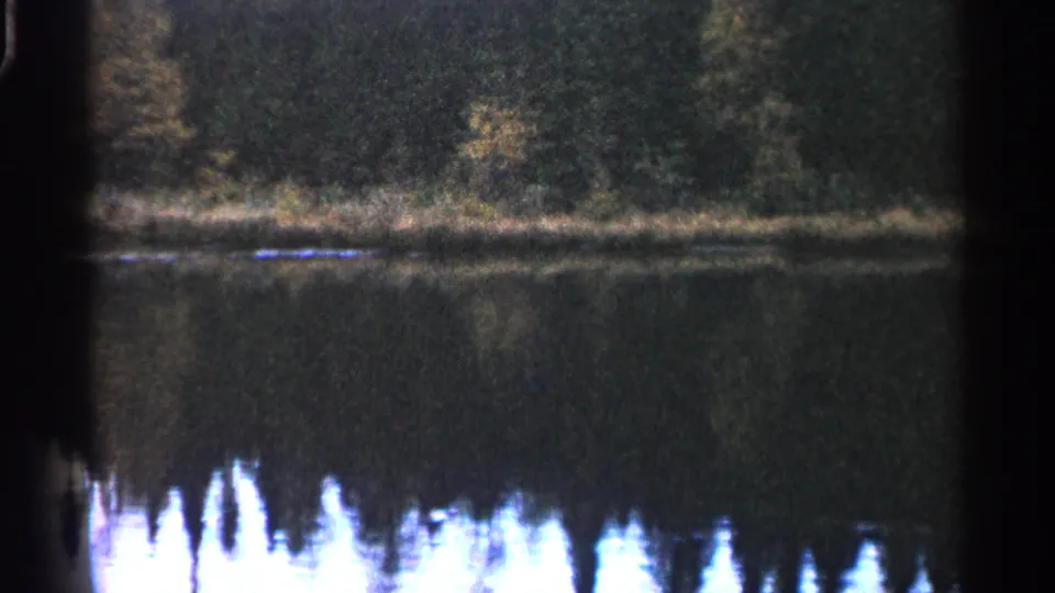 1960 Michigan home-movie frame of a quiet lake and treeline, with strong white reflections and visible 8mm grain.