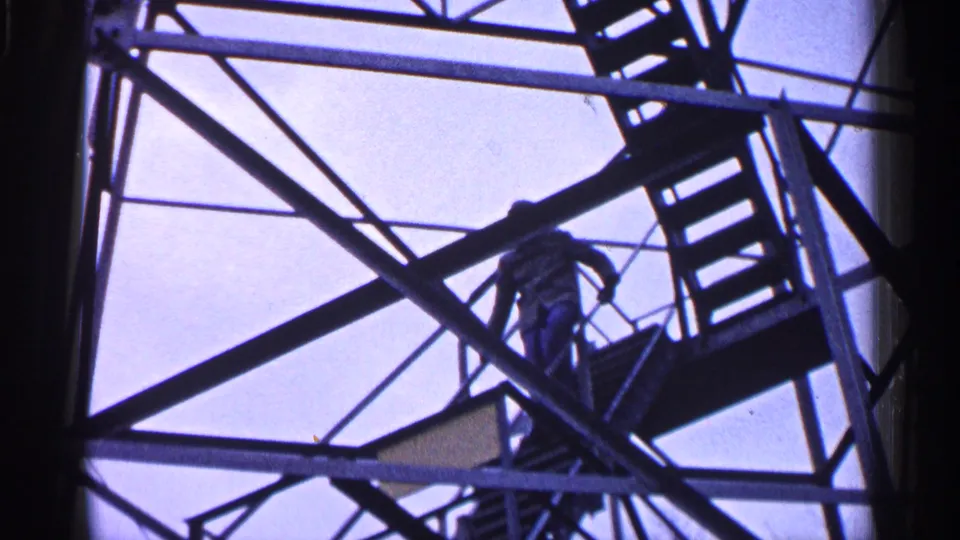 Silhouetted person on a metal stair or tower structure, seen from below in 1960 Michigan 8mm home-movie footage.