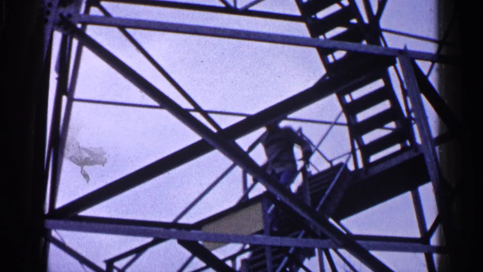 1960 Michigan 8mm frame of a man in a striped shirt climbing stairs inside a steel tower framework, viewed from below.
