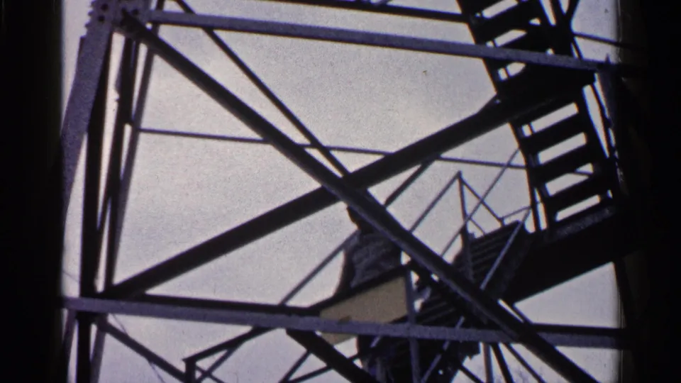 A low-angle 8mm view shows the man on the staircase inside the tower, with heavy diagonal beams dominating the frame.