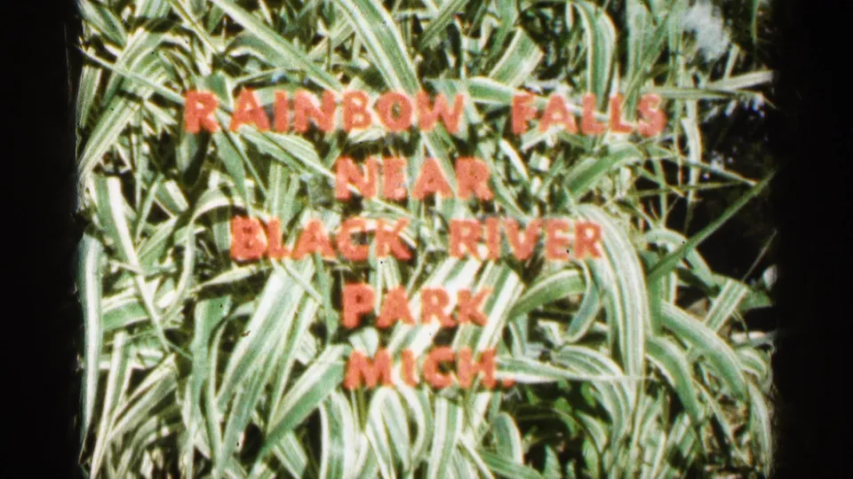 1960 home-movie title card with red lettering over leafy plants identifying Rainbow Falls near Black River Park in Michigan.