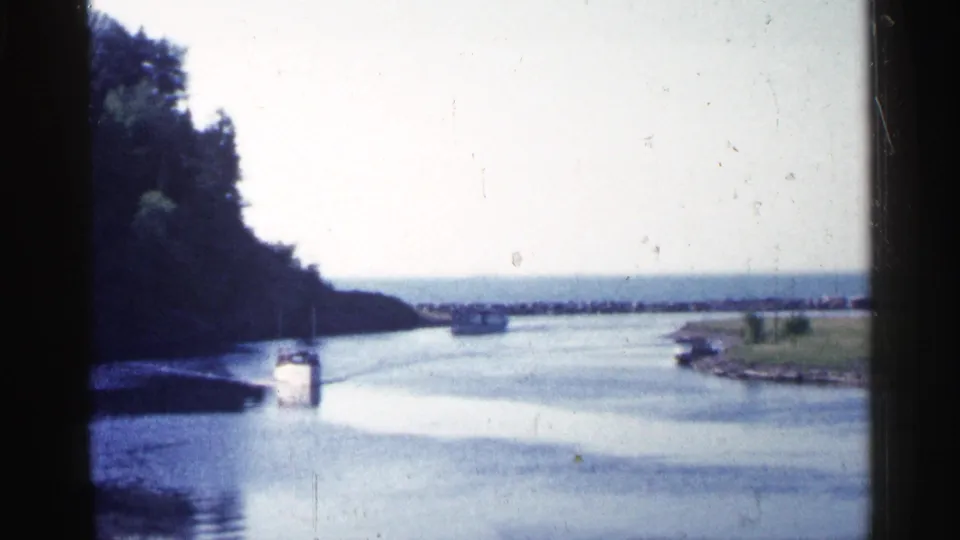 1960 Michigan river view with small boats on calm water, wooded shoreline, pale sky, and distant open water beyond.