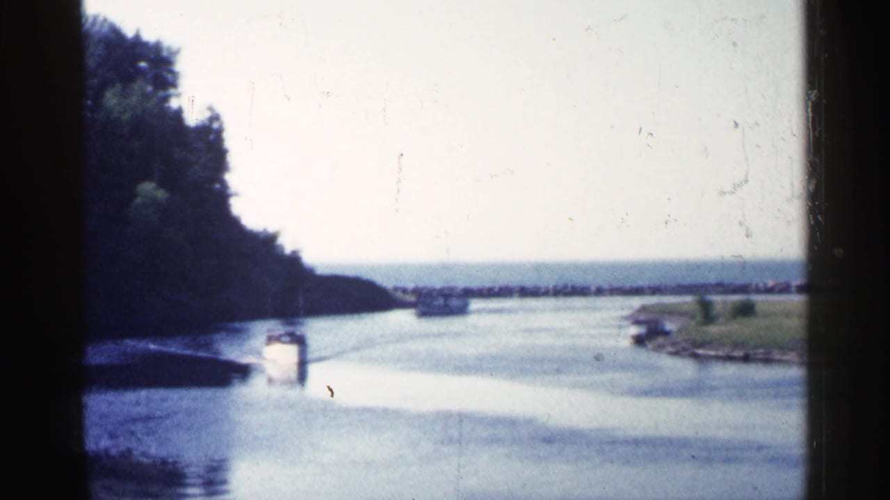 1960 Michigan 8mm frame showing a small white boat on a river channel, with trees at left, grass at right, and dark film edges.