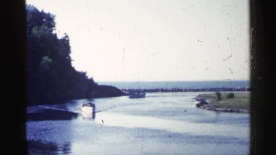 1960 Michigan 8mm frame showing a small white boat on a river channel, with trees at left, grass at right, and dark film edges.