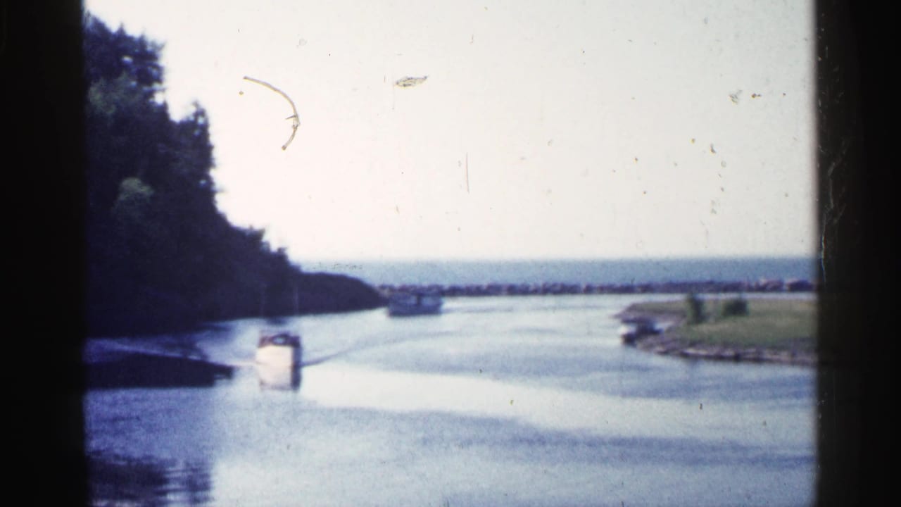 The boat continues through the Michigan river channel, framed by a dark tree line, grassy shore, pale sky, and visible film gate edges.