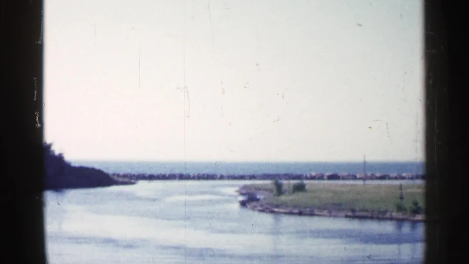 Wide 8mm view of a Michigan river bend, with a small boat near the grassy bank and a low breakwater line across the distant water.