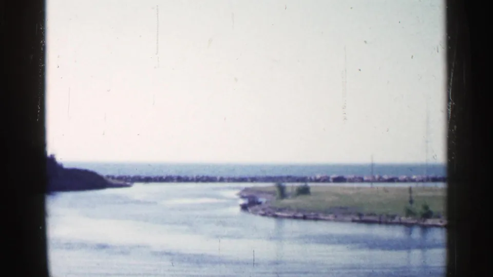 Final 1960 Michigan frame showing the boat near the grassy shoreline, broad river surface, distant breakwater, and soft faded 8mm color.