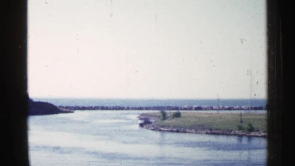1960 Michigan home-movie frame showing a still lake or harbor channel, grassy point, and distant breakwater through a window frame.