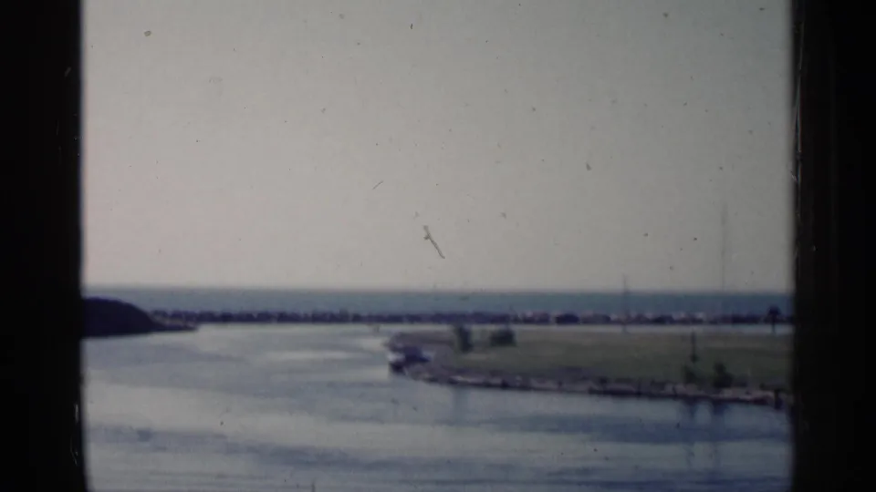 1960 Michigan lake or harbor view with a calm inlet, grassy point, low breakwater, and faint mast-like shapes on the horizon.