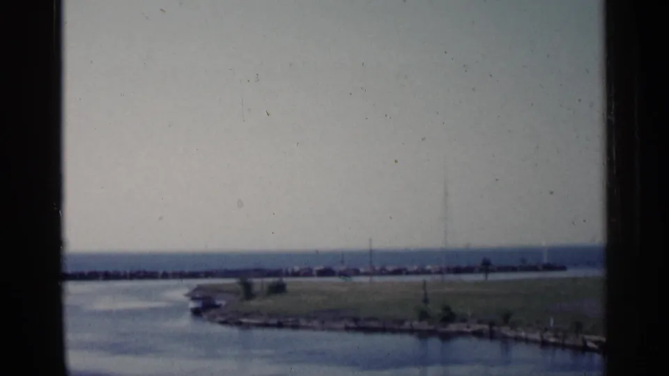 1960 Michigan waterfront frame with a low grassy shore, calm blue water, and dark breakwater line in soft home-movie focus.
