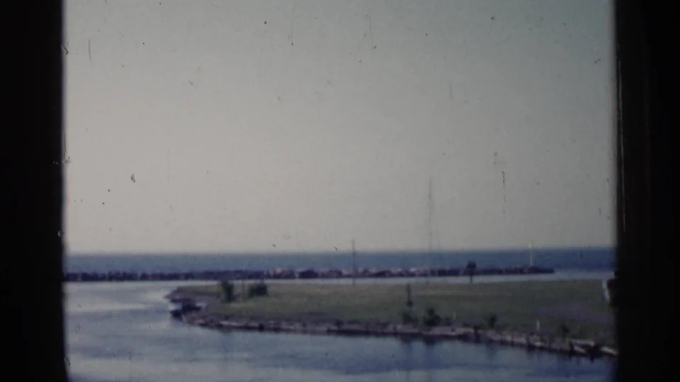 1960 Michigan home-movie still showing a grassy peninsula, calm water, distant breakwater, and faint vertical posts or masts.
