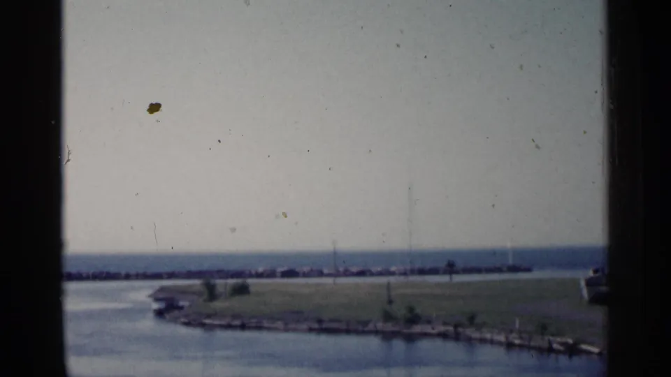 1960 Michigan 8mm frame showing a calm shoreline through window trim, with a grassy point and distant breakwater under hazy sky.