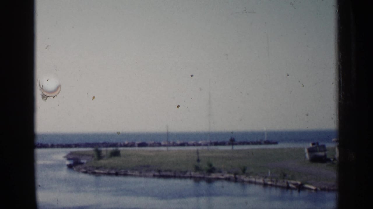 Waterfront scene with window mullions, grassy point, distant breakwater, and small boat at right.