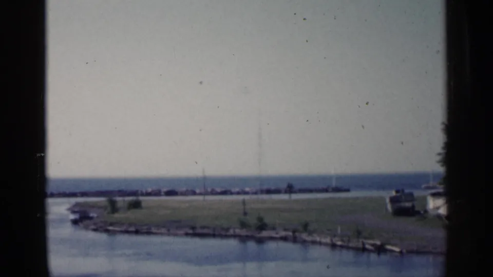 Muted 1960 Michigan 8mm frame of a lake and grassy shore, with a breakwater across the distance and a boat partly visible near the right side.