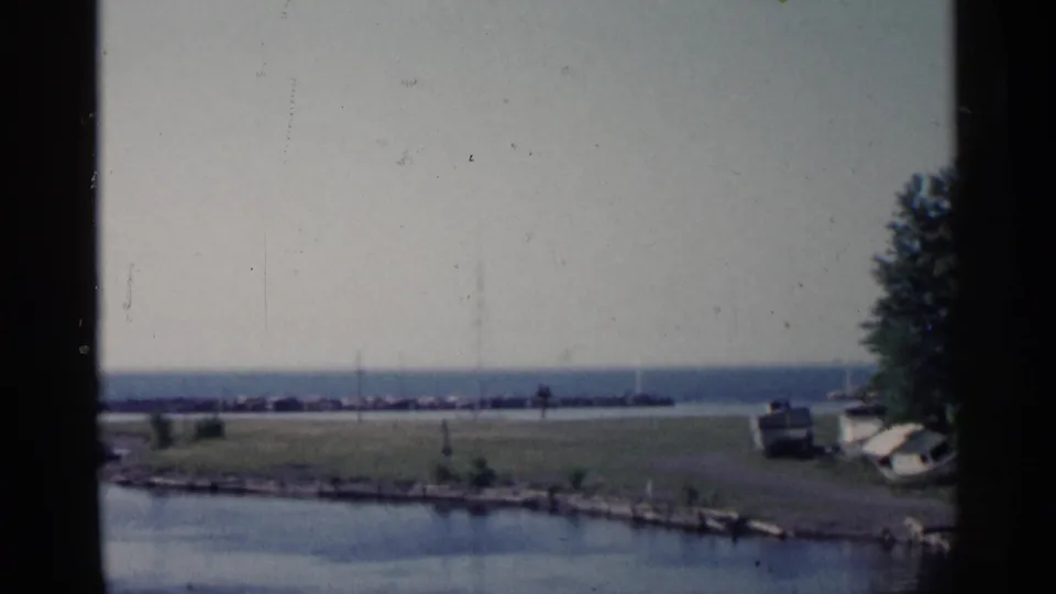1960 Michigan home-movie frame showing a still lakeside scene through window trim, with a boat, trees at right, water, and distant breakwater.