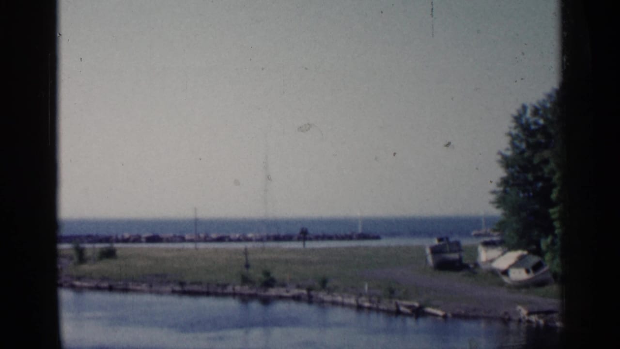 1960 Michigan 8mm frame showing a quiet harbor shoreline with calm water, parked cars, trees, and a breakwater under a pale sky.