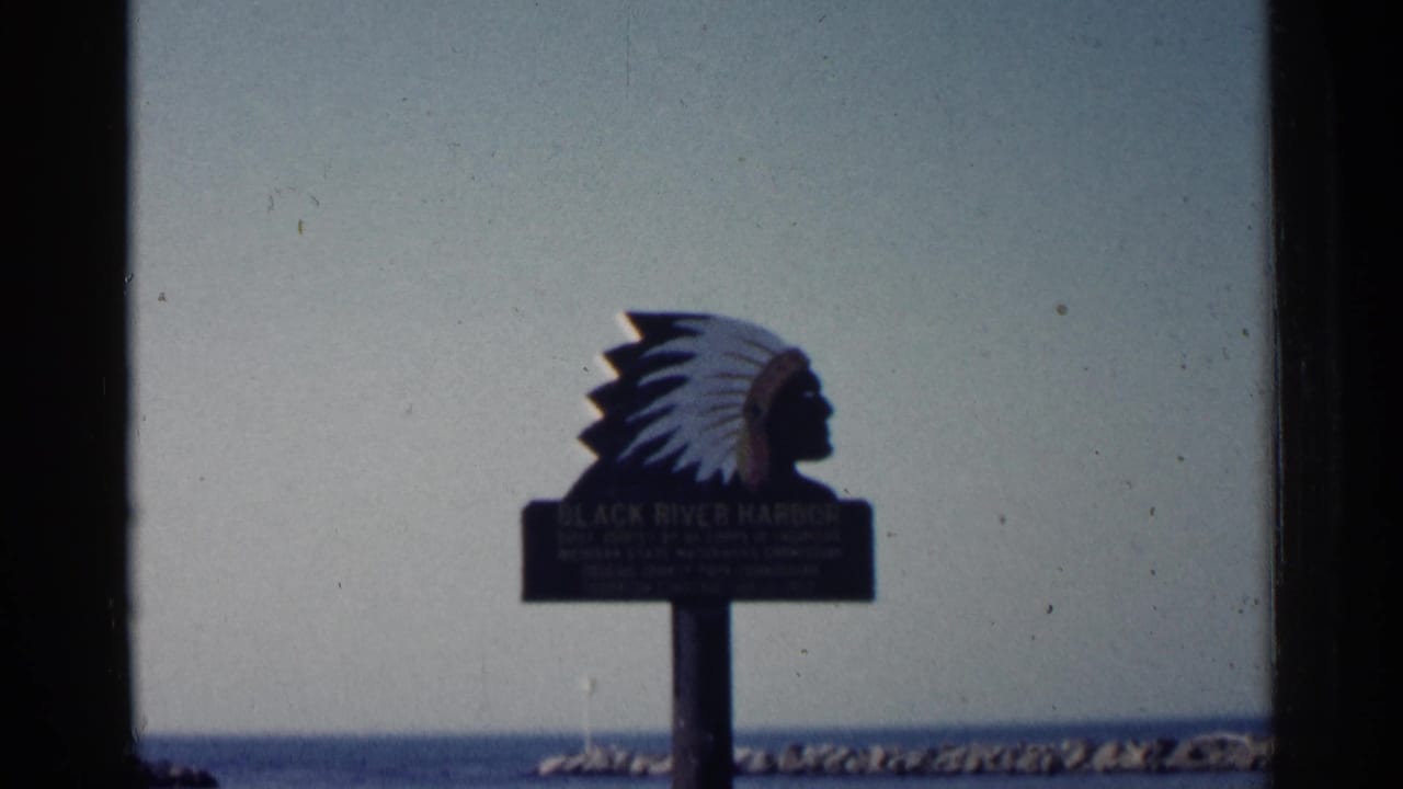Black River Harbor sign topped by a Native American head profile, with lake and breakwater behind it.