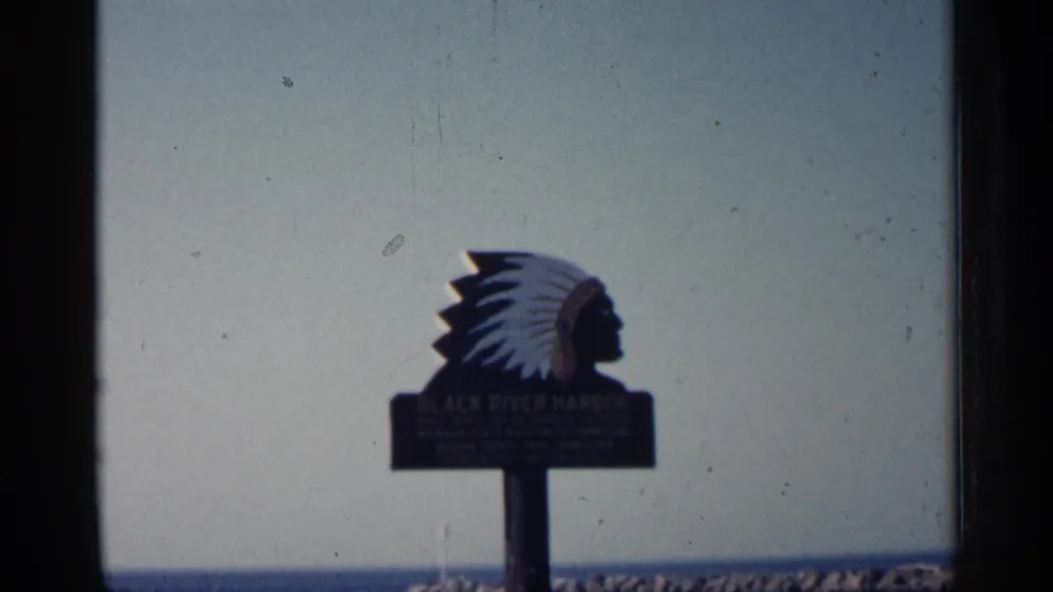 Visitor-shot 1960 Michigan frame of the Black River Harbor sign with a Native American head silhouette above the lake and rock breakwater.