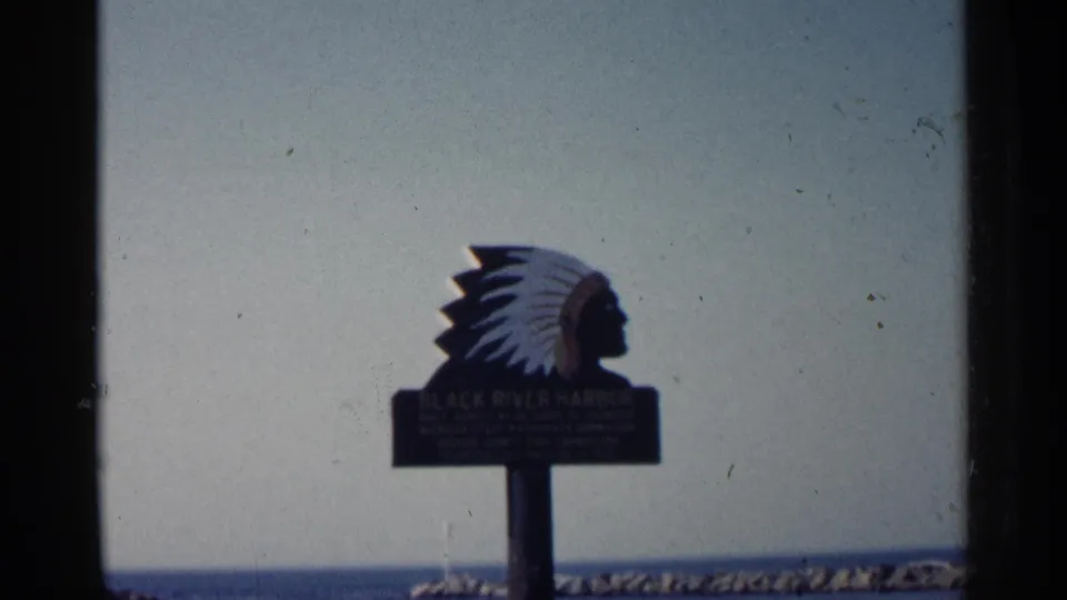 1960 Michigan 8mm frame holding on the Black River Harbor sign, with faded color, visible gate edges, lake, and breakwater behind it.
