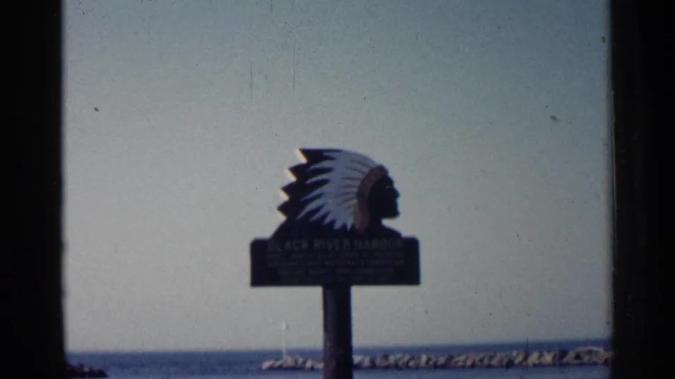 1960 Michigan home-movie frame showing the Black River Harbor sign with Native American chief silhouette, water, pale sky, and breakwater.