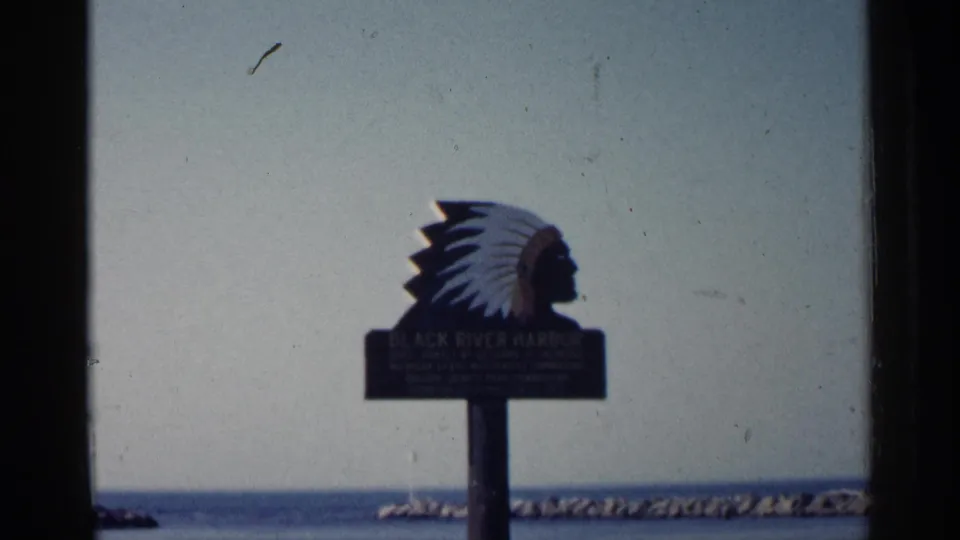 1960 Michigan frame with the Black River Harbor sign and Native American chief silhouette, viewed across water and a low breakwater.
