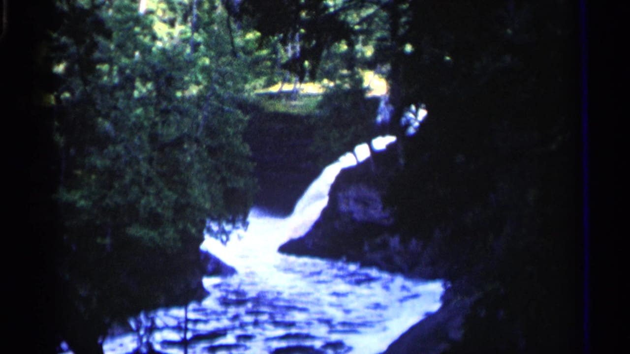 1960 Michigan 8mm frame of a small waterfall dropping into a dark rocky river channel surrounded by leafy trees.