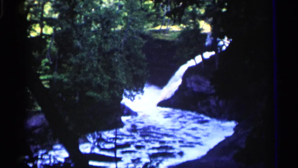 1960 Michigan 8mm frame looking through trees toward a bright waterfall entering a rocky river below.