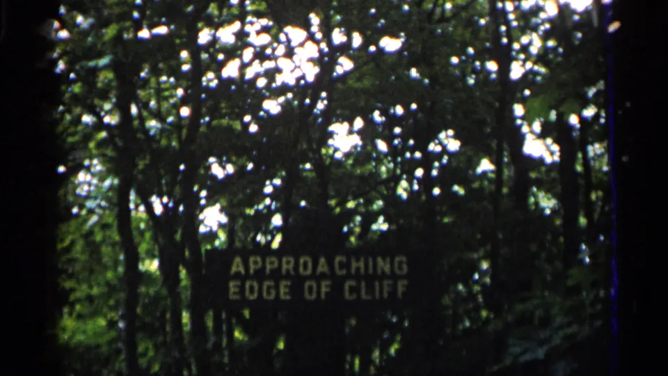 1960 Michigan 8mm frame showing a dark trail sign reading APPROACHING EDGE OF CLIFF in a wooded area.