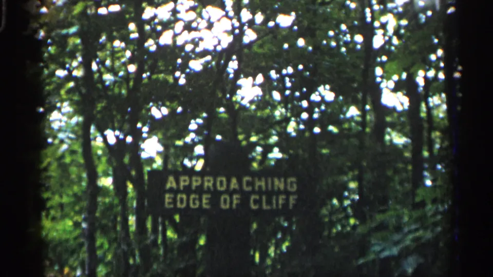 1960 Michigan 8mm frame of an APPROACHING EDGE OF CLIFF sign standing among trees and filtered sunlight.