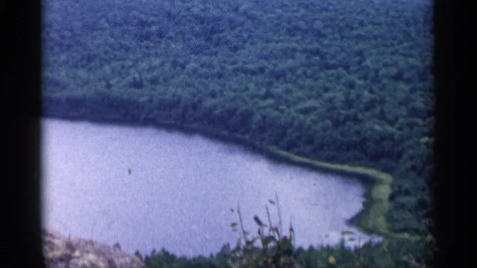 High-angle 1960 Michigan 8mm view of a blue lake, wooded shoreline, and rocky foreground from a scenic overlook.