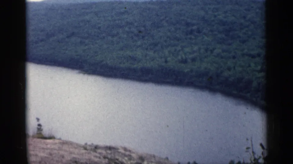 1960 Michigan 8mm frame showing a broad river curve beneath wooded hills from a rocky hillside overlook.