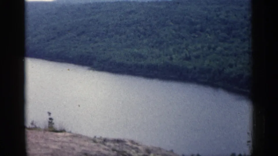 Soft 8mm view of a Michigan river valley with wooded slopes, pale water, and a bare hillside in the foreground.