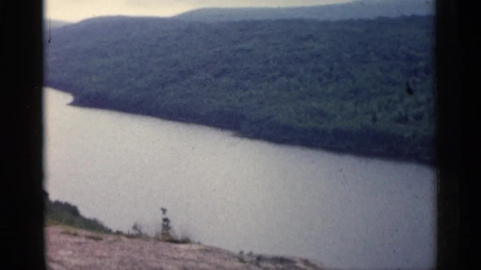 1960 Michigan home-movie frame showing a curving river at the base of forested hills, filmed from a scenic overlook.