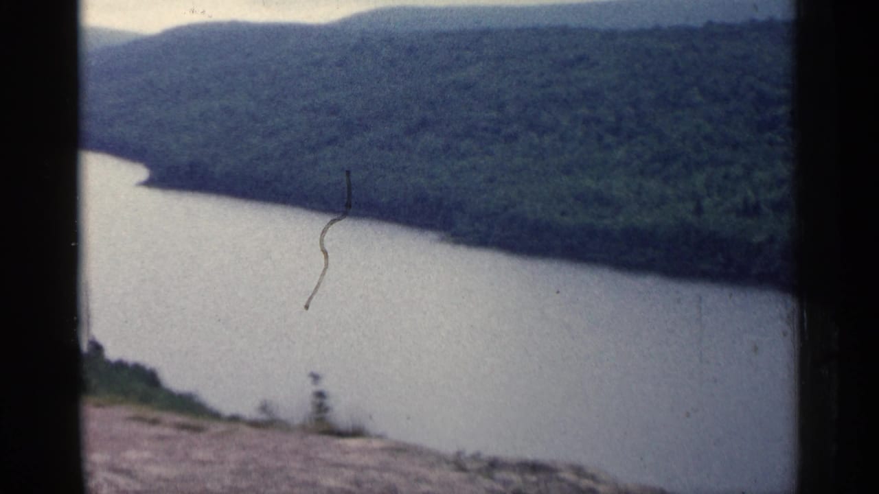 1960 Michigan 8mm view of a broad river curving below wooded hills, with rocky foreground and visible film gate edges.