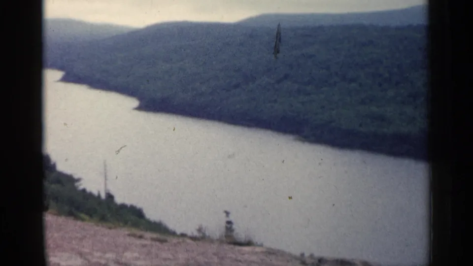 1960 Michigan 8mm frame of a river valley from an overlook, with muted color, rocky foreground, and forested hills across the water.