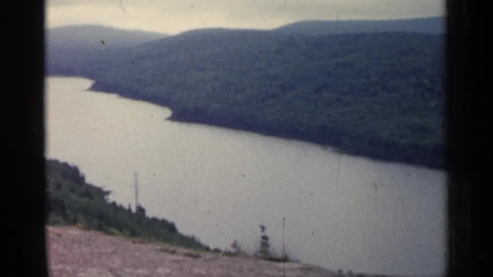 1960 Michigan 8mm view across a river and forested valley, with subdued exposure, visible grain, and no people or buildings apparent.