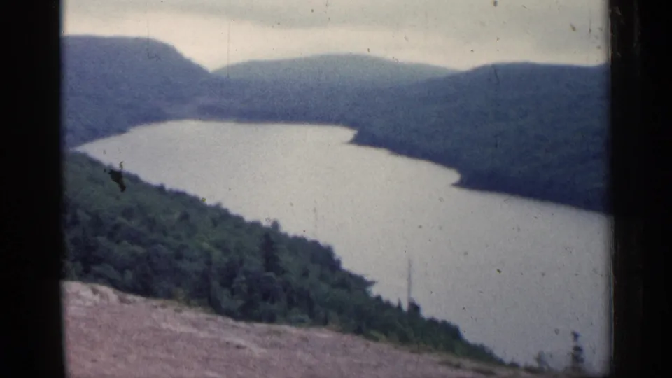 1960 Michigan 8mm frame showing a broad bend of water enclosed by forested hills beneath cloud cover.