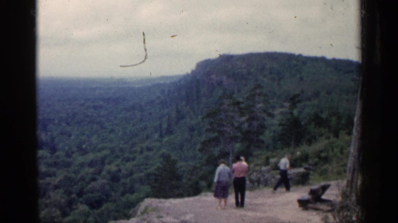 1960 Michigan 8mm frame showing three people at a cliffside overlook above a wooded valley and hazy ridge.