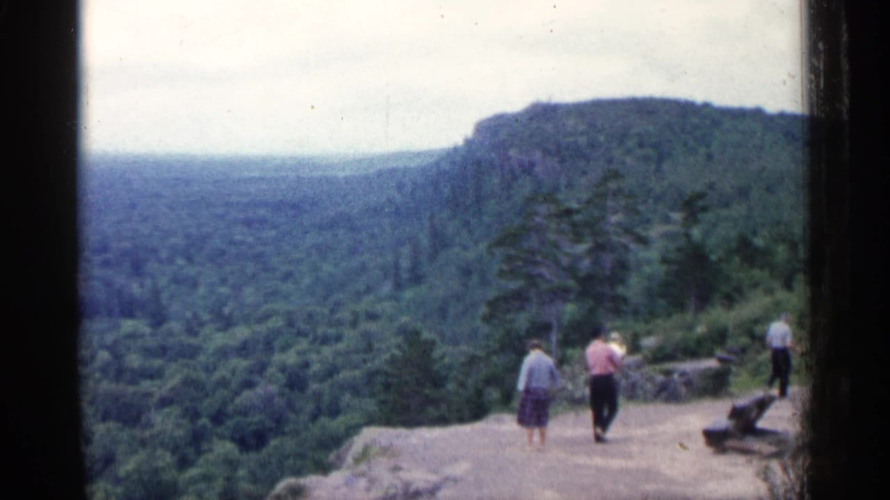 1960 Michigan home-movie view of visitors near a rocky scenic overlook with forest and ridge beyond.