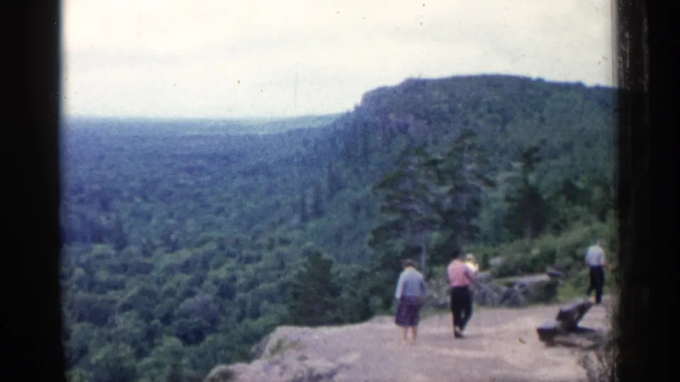 1960 Michigan home-movie view of visitors near a rocky scenic overlook with forest and ridge beyond.