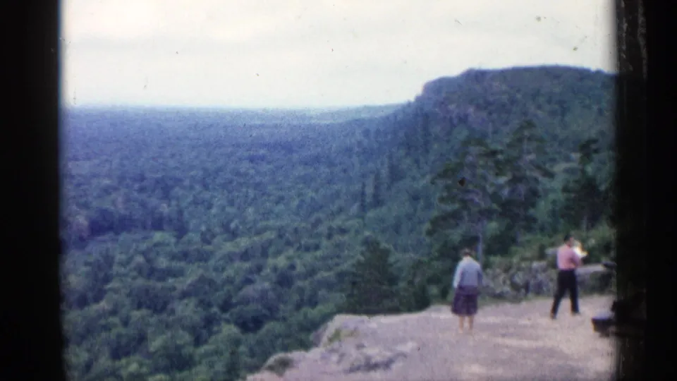 1960 Michigan 8mm frame with two visitors at a cliff overlook facing a wide forested valley.
