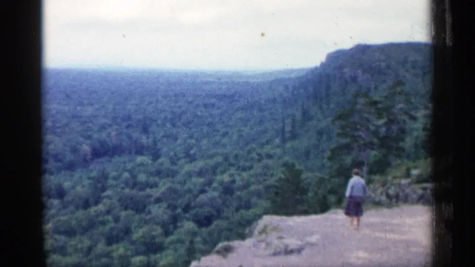 1960 Michigan home-movie frame of a lone visitor on a rocky overlook above wooded hills.