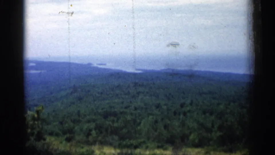A soft 1960 Michigan 8mm frame shows a broad forest canopy below distant lake water and a bright clouded sky.